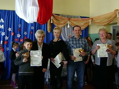 Grupa dorosłych z dyplomami i kwiatami na szkolnej uroczystości.