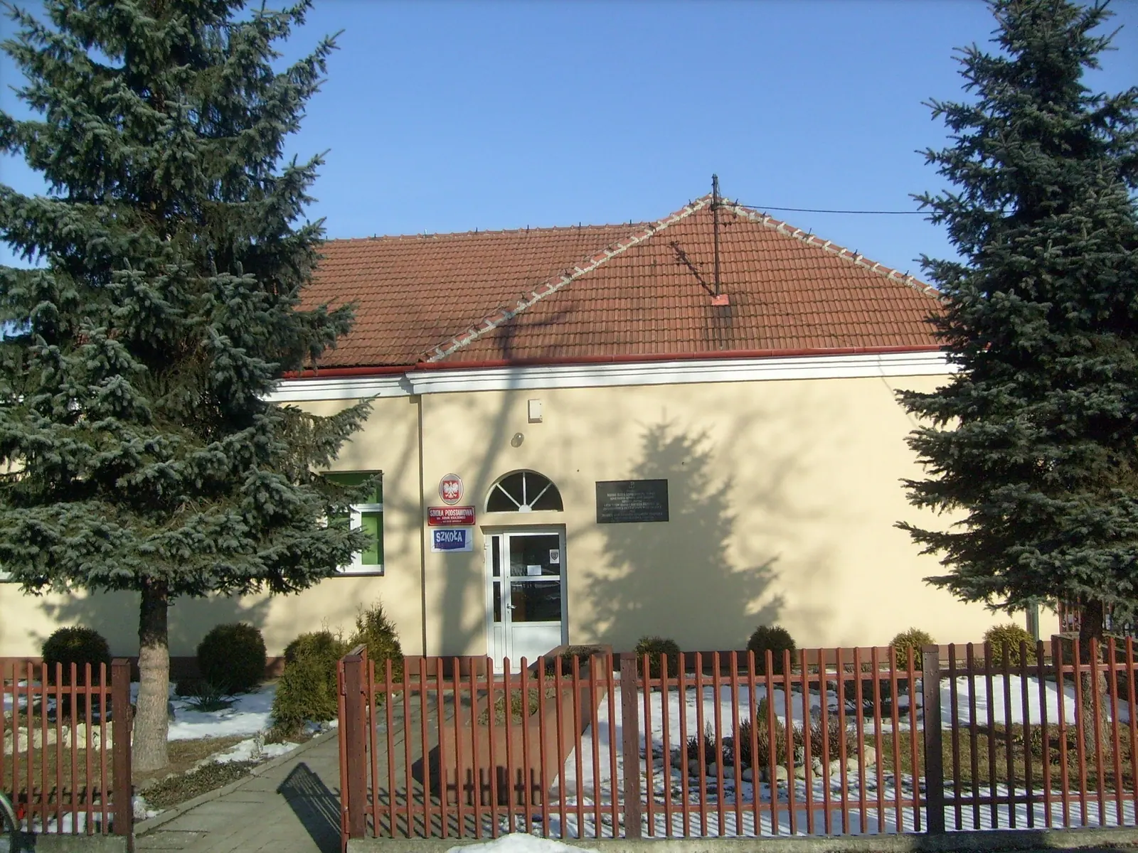Jasnożółty parterowy budynek szkoły z czerwonym dachem, przed nim ogrodzenie i zieleń.
