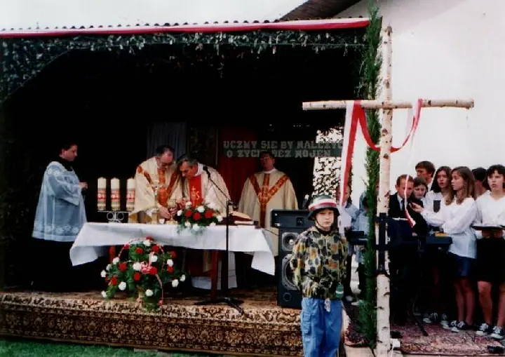 Uroczystość przed budynkiem szkoły: poczty sztandarowe, flagi biało-czerwone, mieszkańcy i kombatanci.