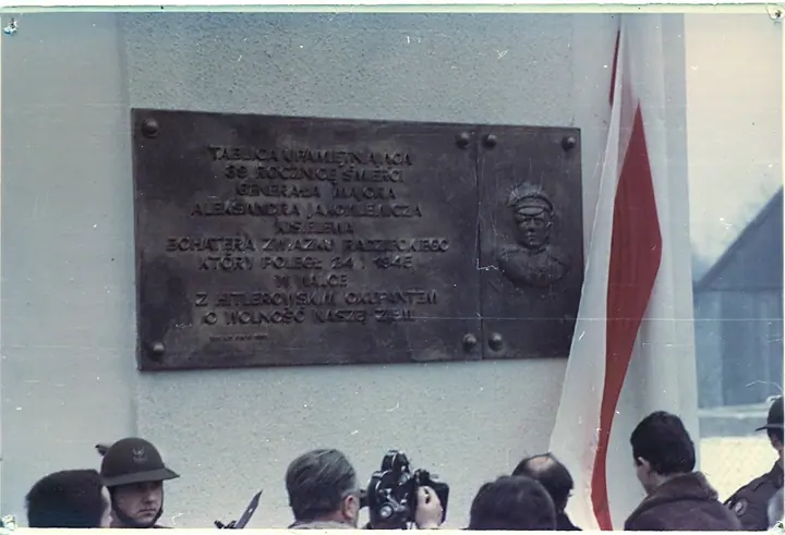 Ciemna tablica pamiątkowa na ścianie szkoły, obok flaga i grupa uczestników uroczystości.