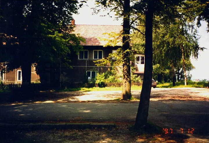 Parterowy budynek wikarówki wśród wysokich drzew.