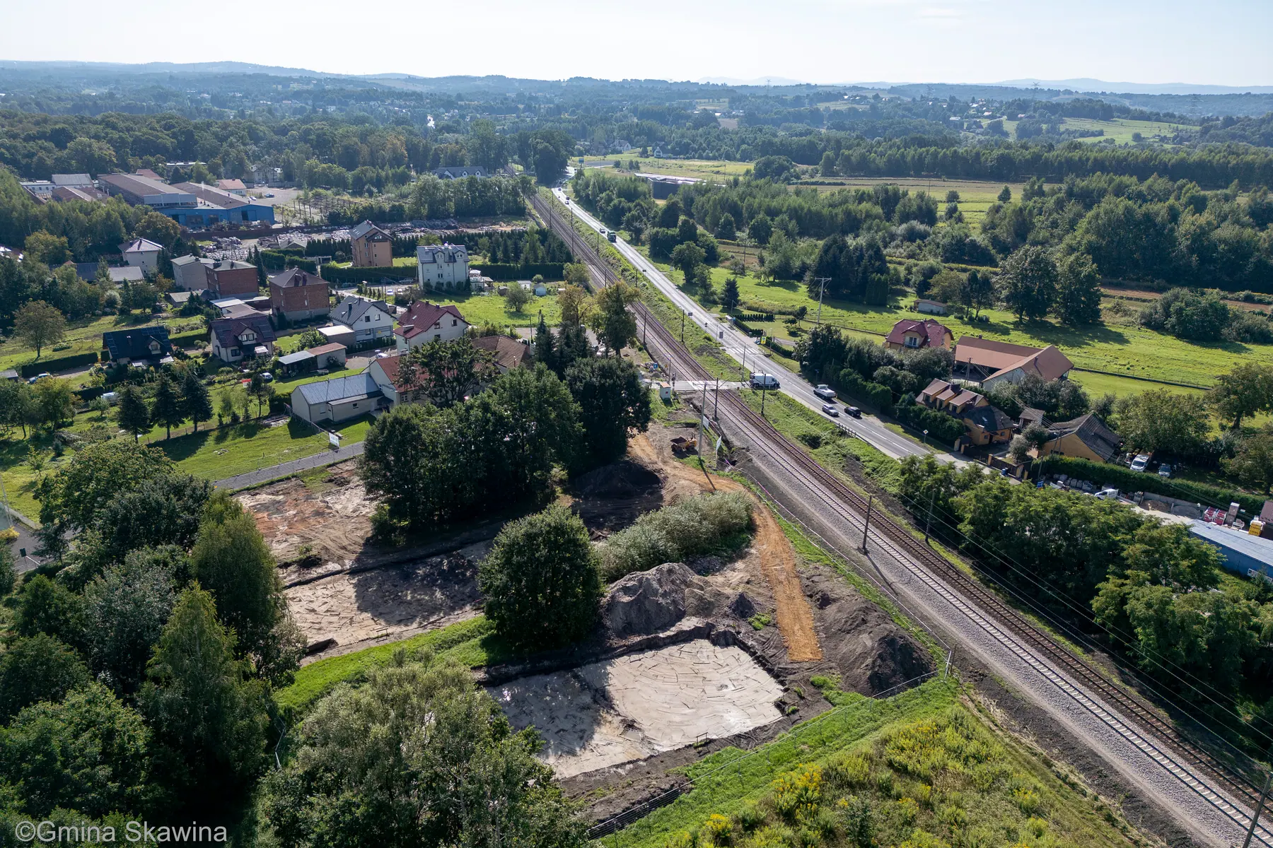 Widok z drona wzdłuż torów kolejowych na oczyszczony plac budowy pośród zieleni.