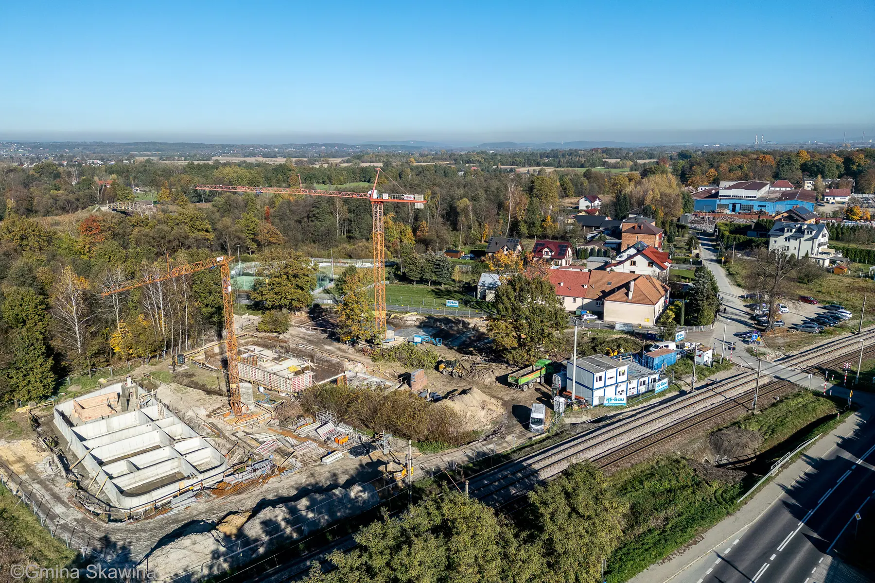 Plac budowy z betonowymi płytami fundamentowymi i żurawiem, w tle zabudowa wsi.