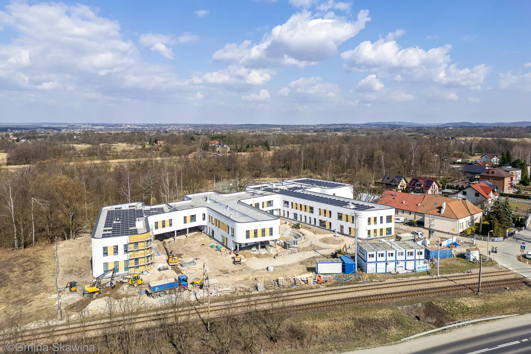 Biało-musztardowe budynki kompleksu, wokół nich niewykończony teren i sprzęt budowlany.